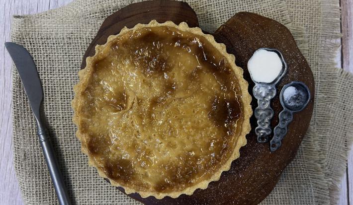 Foto vista de cima da receita de Torta de Leite Moça, sobre uma tábua de madeira, com duas colheres de ferro ao lado, uma delas com açúcar. Abaixo da tábua há um tecido e sobre ele uma faca do lado esquerdo.
