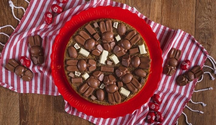 Fotografia em tons de vermelho, em uma bancada de madeira de cor marrom. Ao centro, uma toalha vermelha listrada com um prato vermelho em cima contendo o cookie em fatias. Ao redor do prato, há alguns KitKat's bunny e mini ovinhos de KitKat.
