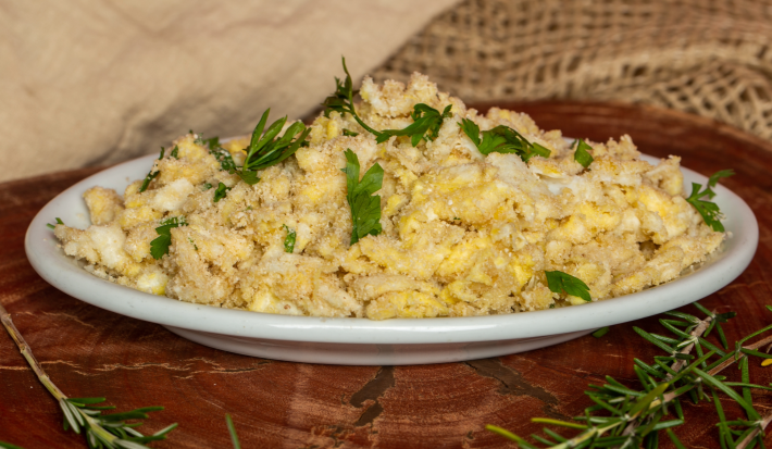 Fotografia em tons de amarelo com um prato branco ao centro. Em cima do prato existe uma receita de farofa de ovos feita com farinha de mandioca e finalizada com cheiro-verde.