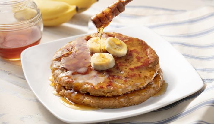 Fotografia me tons de branco de uma bancada de madeira, sobre ele um paninho listrado azul e branco com um prato quadrado com duas panquecas, mel e bananas. Ao fundo duas bananas e um recipiente de vidro com mel.