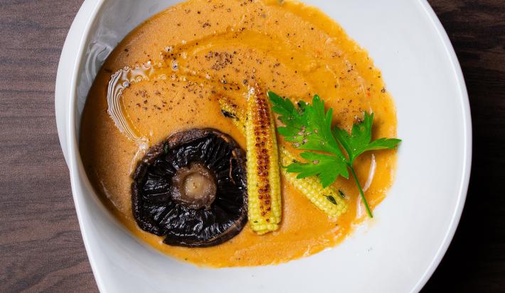 Fotografia em tons de branco com um prato branco ao centro. Em cima do prato existe uma receita de sopa de cor alaranjada com pedaços de cogumelo, mini milho e coentro em cima.