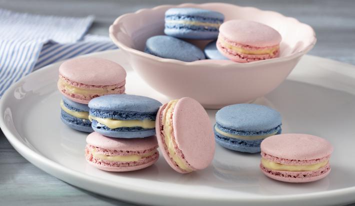 Fotografia em tons de azul e rosa em uma bancada de madeira branca clara com vários macarons coloridos em um prato branco e um paninho azul listrado.