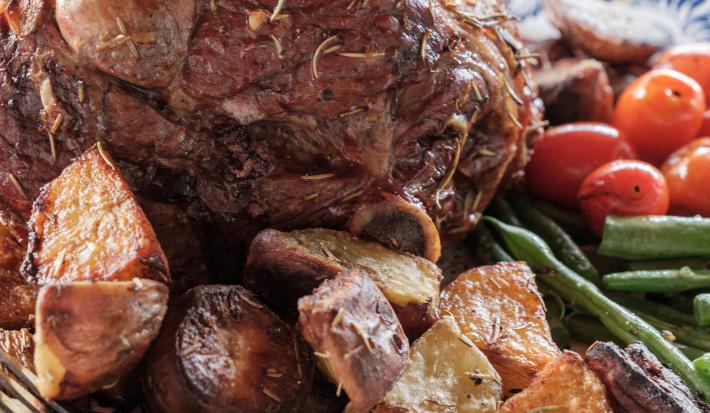 Fotografia de um cordeiro assado com ervas, batatas assadas, vagens e tomates cereja.