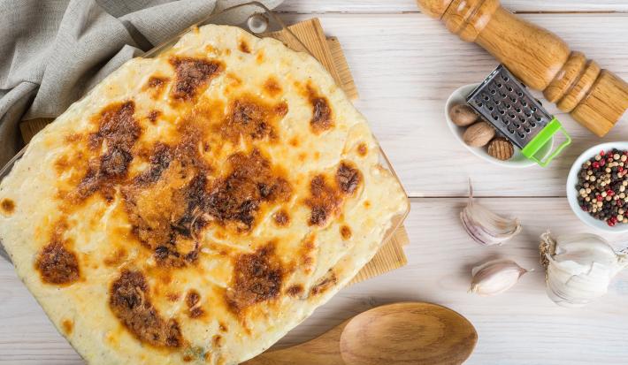 Fotografia de uma travessa de vidro quadrada de bacalhau gratinado com batata sobre uma tábua de madeira e um pano de tom cinza. No fundo tem um moedor de pimenta de madeira.