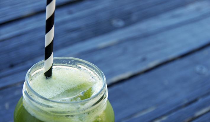 Fotografia de uma mesa de madeira preta, sobre ela tem um copo grande decorativo de vidro com um suco de Kiwi, abacaxi, chia e hortelã. Dentro do copo tem gelo e um canudo preto e branco.
