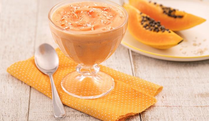 Foto em tons de laranja da receita de creme de papaia servida em uma taça de vidro sobre um pano laranja em uma mesa de madeira. Ao lado há um colher prateada e ao fundo há um prato com duas fatias médias de mamão