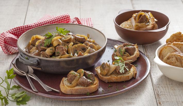 Fotografia em tons de marrom em uma bancada de madeira clara com um pano vermelho xadrez, um prato redondo marrom grande com pães e carnes em cubos e ao centro uma panelinha marrom com mais carne e cogumelo. Ao lado, um potinho com pães.
