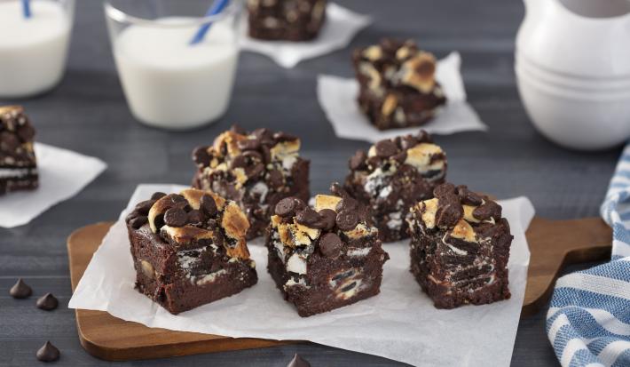 Foto da receita de Brownie de Negresco Coberto, em pedaços, sobre uma tábua de madeira em um balcão decorado com alguns copos com leite, um tecido azul listrado e algumas gotas de chocolate