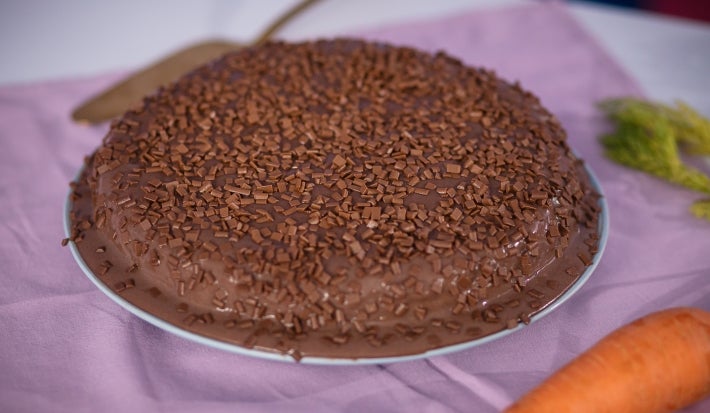 Fotografia de um bolo de cenoura com cobertura de chocolate e ascenda sobre um prato raso grande, apoiado em um pano roxo quadrado.