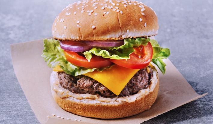 Fotografia com um fundo cinza, um pedaço de papel manteiga cortado em quadrado. Sobre ele, um hambúrguer com carne bovina, uma fatia de queijo, alface, duas rodelas de tomate, cebola roxa e um molho na base do lanche.