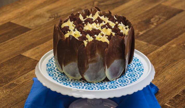 Foto aproximada de um bolo de chocolate com decoração de lascas marmorizadas, sobre um prato branco e azul em uma bancada de madeira