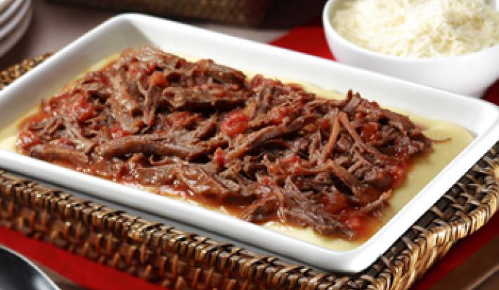Fotografia em tons de vermelho, branco e marrom, com refratário sobre bandeja de vime com carne desfiada e molho de tomate, sobre guardanapo vermelho, ladeada por pote branco com arroz e uma colher, colher de servir, pratos, tudo sobre bancada de madeira.