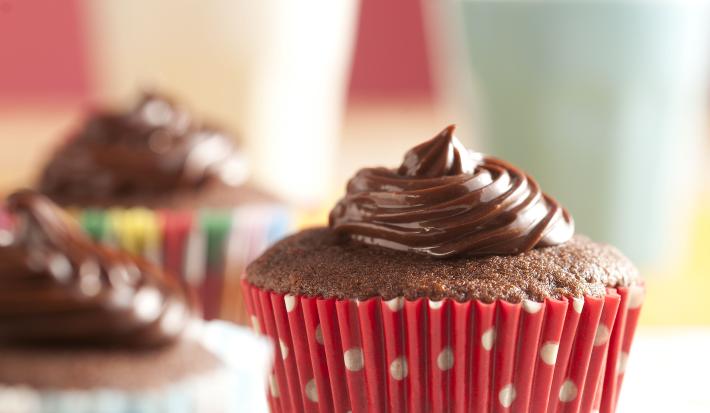 Fotografia em tons de branco e vermelho de uma bancada branca com bolinhas coloridas, sobre ela três cupcakes de Nescau. Ao fundo dois copos com canudos coloridos.