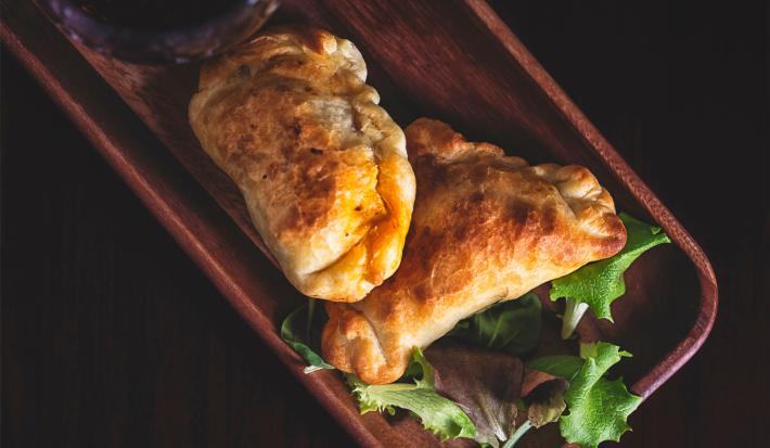 Fotografia em tons escuros vista de cima de duas empanadas recheadas com sardinha sobrepostas e apoiadas em uma tábua de madeira retangular escura.