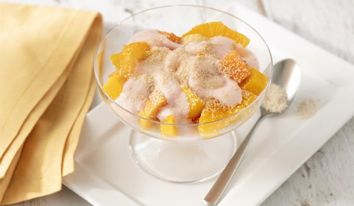 Fotografia em tons de branco e amarelo de uma bancada branca com um prato retangular branco, sobre ele uma taça com a salada de frutas e uma colher. Ao lado um paninho amarelo.