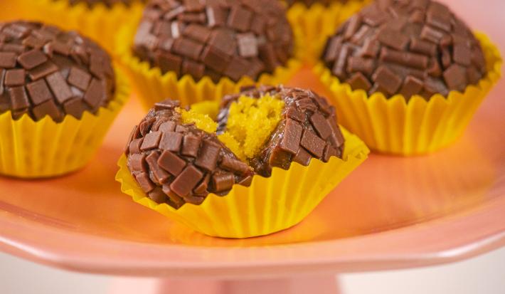 Fotografia em tons de amarelo, rosa e marrom. Ao centro um brigadeiro mordido, mostrando o recheio de bolo de cenoura.
