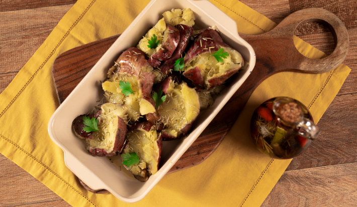 Fotografia em tons de amarelo com uma tábua de madeira ao centro. Em cima da tábua existe uma travessa clara com batatas doces ao murro gratinadas e temperadas com alecrim e azeite