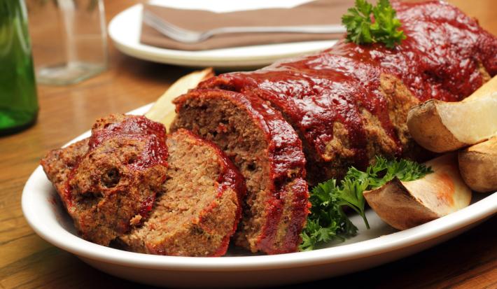 Fotografia de carne com aveia ao forno, metade cortada em fatias e dourada por cima dentro de um refratário branco oval. Ao lado, algumas batatas assadas, e ao fundo, um prato branco com um garfo encima, sobre uma mesa de madeira.