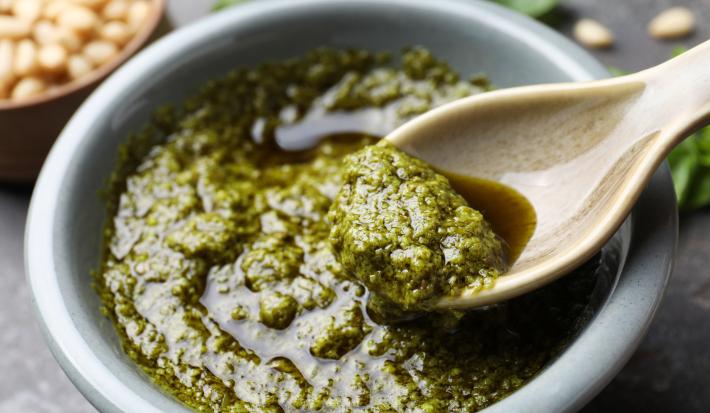 Foto aproximada da receita de molho pesto sem oleaginosas, servido em uma tigela redonda em que há uma colher pegando uma pequena quantidade dele