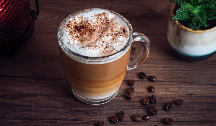 fotografia em tons de marrom e branco de uma bancada marrom vista de cima. Contém uma xicara transparente com café e por cima chantilly com raspas de chocolate.