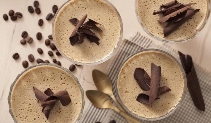 Fotografia em tons de branco e marrom de uma bancada branca, sobre ela grãos de café, um paninho branco e preto e quatro taças com mousse de café decorados com chocolate.