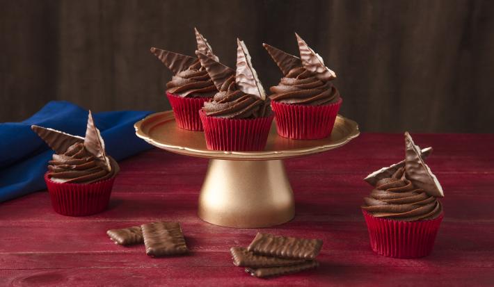 Foto de uma bancada vermelha. Ao centro há um suporte para bolos dourado alto com 3 cupcakes em formas vermelhas e decorados com Nestlé ChocoBiscuits. Na bancada há mais 2 cupcakes, alguns biscoitos e um tecido azul escuro ao fundo.