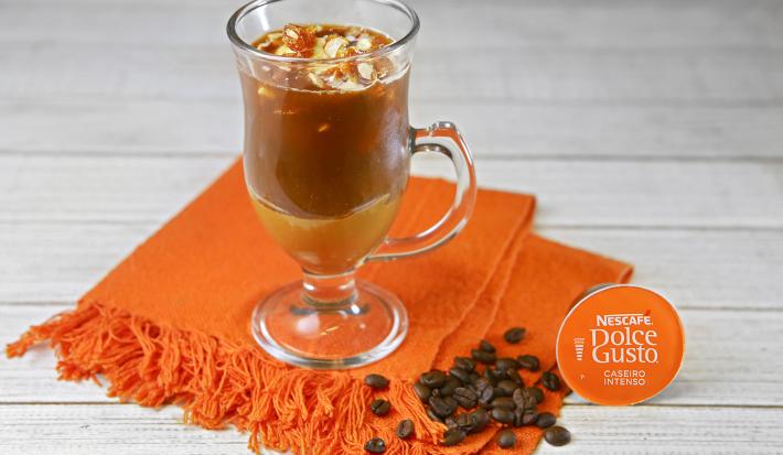 Fotografia em tons de laranja em uma bancada de madeira branca, um pano laranja, uma xícara de vidro com o café com sorvete e pé de moleque dentro dele. Ao lado, uma cápsula de café caseiro intenso Dolce Gusto. Fotografia em tons de laranja em uma bancada de madeira branca, um pano laranja, uma xícara de vidro com o café com sorvete e pé de moleque dentro dele. Ao lado, uma cápsula de café caseiro intenso Dolce Gusto.