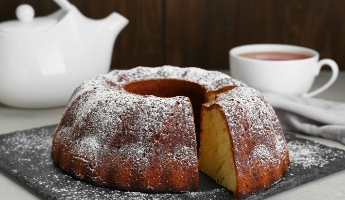 Fotografia de um bolo bundt recém-assado em uma tábua escura, polvilhado com açúcar de confeiteiro. Um bule branco e uma xícara de chá estão ao fundo.