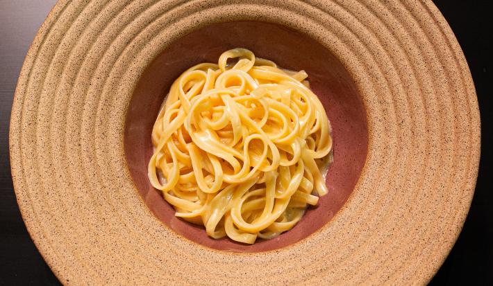Fotografia em tons de cinza vermelho com um prato vermelho claro ao centro. Dentro do prato existe uma receita de fettuccine Alfredo feita com manteiga e queijo.
