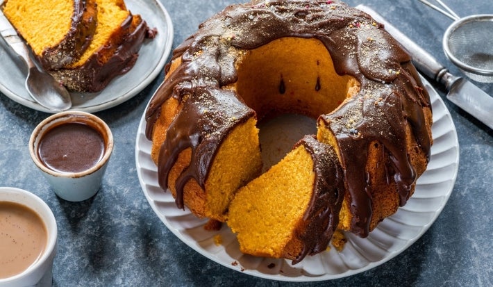 Bolo de cenoura com cobertura de chocolate em prato branco. Uma fatia retirada mostra o interior macio e amarelo. Ao lado, xícaras com calda e café, e um prato com fatias servidas. A cobertura de chocolate escorre generosamente pelas laterais.
