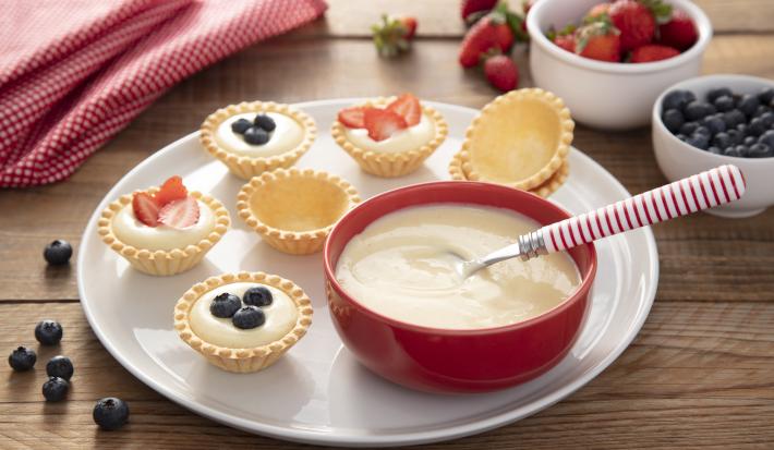 Fotografia em tons de vermelho e amarelo em uma bancada de madeira, um pano vermelho de bolinhas, um prato branco redondo com várias tortinhas recheadas com creme de confeiteiro e frutinhas. Ao lado, um potinho vermelho com o creme de confeiteiro.