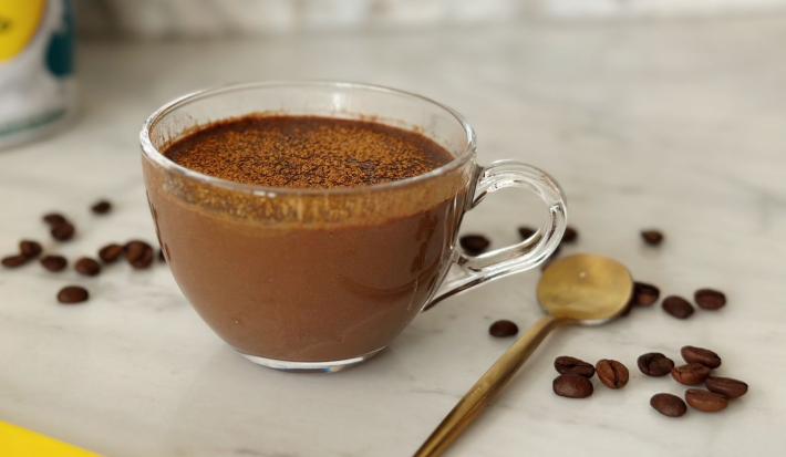Foto aproximada de uma receita de café cremoso servido em uma xícara de vidro, com uma colher dourada ao lado e alguns grãos de café espalhados na mesa