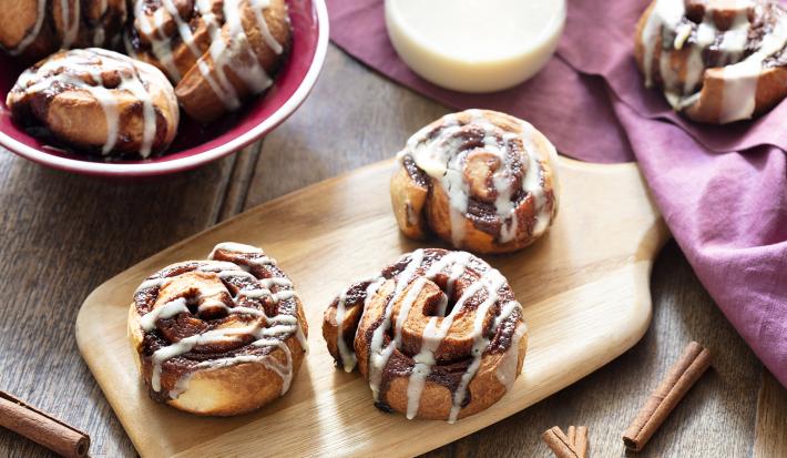 Fotografia em tons de amarelo, em uma bancada de madeira de cor marrom. Ao centro, uma tábua de madeira contendo 3 Cinnamon Rolls. Ao fundo, um pano vermelho e um recipiente contendo cinnamon rolls e uma jarra de leite.