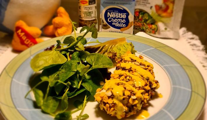 Foto aproximada de um prato redondo com bordas verde e azul com a receita de frango empanado servido com agrião. Ao fundo há produtos Maggi e Nestlé Creme de Leite e uma parte de uma pelúcia da Galinha Azul