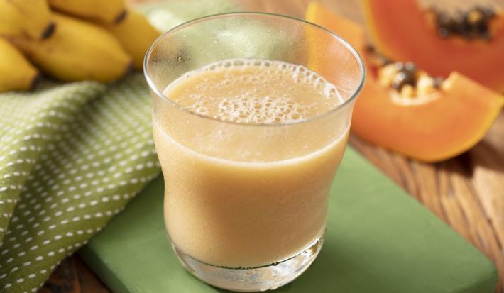 Fotografia em tons de laranja e verde de uma bancada de madeira com um suporte verde e um paninho verde com bolinhas brancas, sobre ele um copo de vidro com a vitamina de mamão e banana. Ao fundo uma fatia de mamão e um cacho de banana.