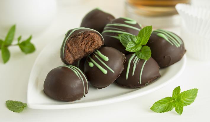 Fotografia em tons de branco, marrom e verde de uma bancada branca vista de frente, contém um prato branco com bombons de chocolate e folhas de hortelã em volta para decorar.