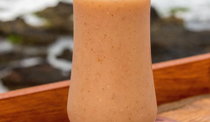 Fotografia de uma taça baixa de vidro com refresco de goiaba com iogurte e NESTON. Dentro da taça tem um canudo de plástico junto com a bebida, que está sobre um apoio de madeira, e ao fundo tem o mar e algumas pedras.