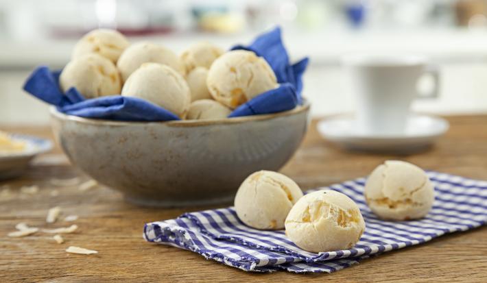 Foto de uma mesa com um pano e 3 pães de queijo redondos em cima. No fundo da foto há uma tigela com mais pães dentro