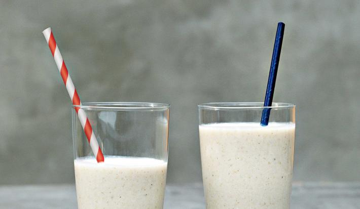 Fotografia em tons de cinza e branco de uma bancada cinza sobre ela dois copos de vidro com vitamina de banana e dois canudos, um azul e um listrado vermelho e branco.
