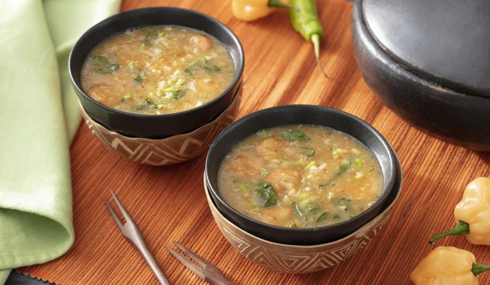 Foto em tons de bege da receita de tacacá servida em duas porções em bowls pretos com adornos em marrom sobre uma esteira de madeira. Ao fundo, há pimentas-de-cheiro e um paninho verde