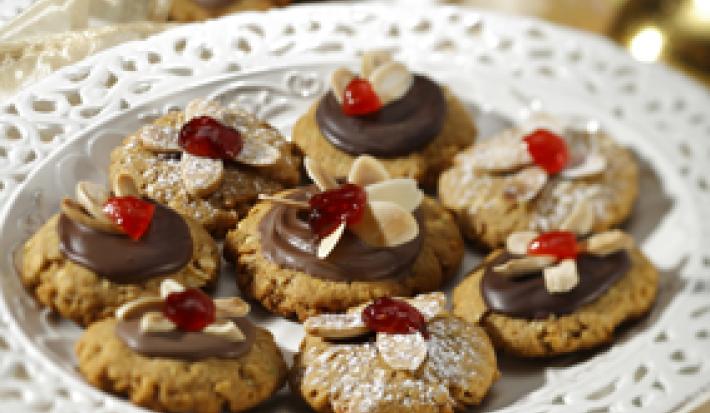 Fotografia em tons de dourado em uma mesa com uma toalha dourada, um suporte para bolo branco com detalhes em dourado e vários biscoitinhos alemães com chocolate e lascas de amêndoas.