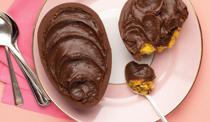 Fotografia em tons de rosa em uma bancada de madeira rosa, um pano rosa, um prato rosa redondo com o ovo de Páscoa de colher de bolo de cenoura recheado com brigadeiro.