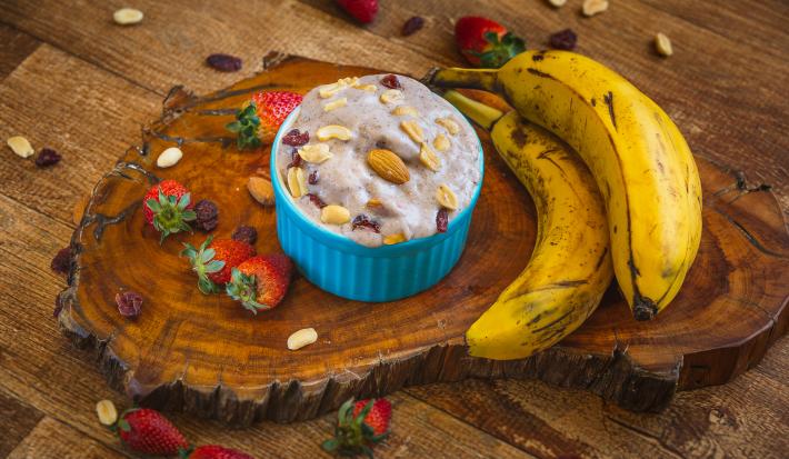 Fotografia em tons de marrom em uma bancada de madeira escura, uma tábua de tronco de árvore, um potinho azul com o sorvete de morango, banana e chia dentro dele e coberto com frutas secas. Ao lado, duas bananas e morangos espalhados pela bancada.