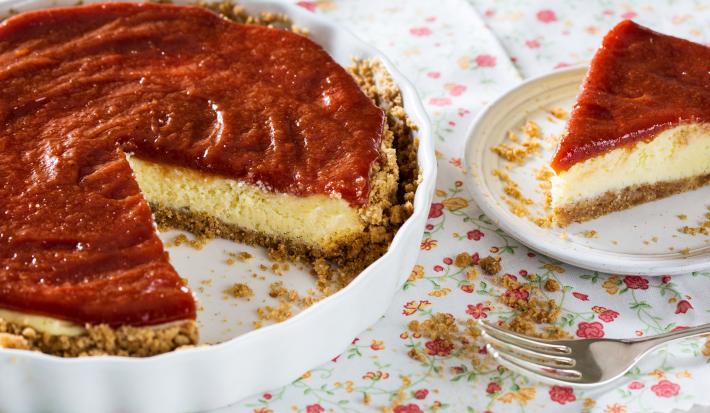 Fotografia em tons de vermelho em uma mesa com uma toalha branca com flores coloridas, um recipiente redondo raso com a cheesecake dentro dele. Ao lado, um prato raso branco com uma fatia da cheesecake.