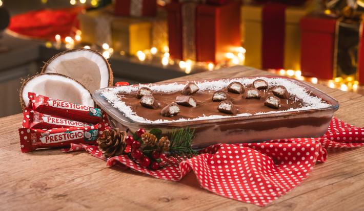 Foto da receita de Pavê de Prestígio. Observa-se o pavê em um recipiente refratário decorado com Prestígios cortados em cima. Atrás, presentes e luzes de Natal decoram a foto.
