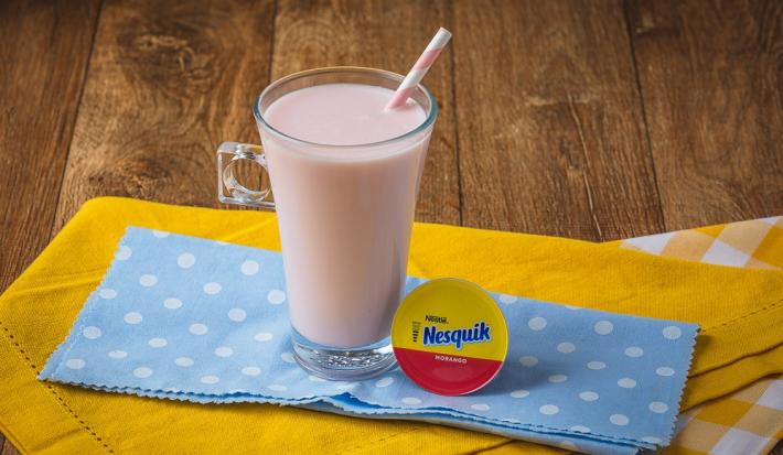 Fotografia em tons de rosa, em uma bancada de madeira de cor marrom. Ao centro, um pano liso amarelo e um pano amarelo quadriculado sobrepostos. Em cima deles, há o frappé de Nesquik com um canudo dentro e ao lado, uma cápsula de Nesquik.