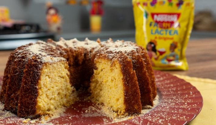 Fotografia de um bolo de milho com farinha láctea apoiado em um prato grande, raso e decorativo de tom vinho. Atrás do bolo tem uma embalagem de farinha láctea.