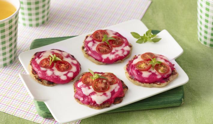 Foto da receita de mini pizza. Observa-se uma bandeja branca com 4 mini pizzas em cima, com recheio de creme rosa de beterraba, tomates cereja e queijo derretido, decoradas com folha de manjericão. O fundo é verde claro.