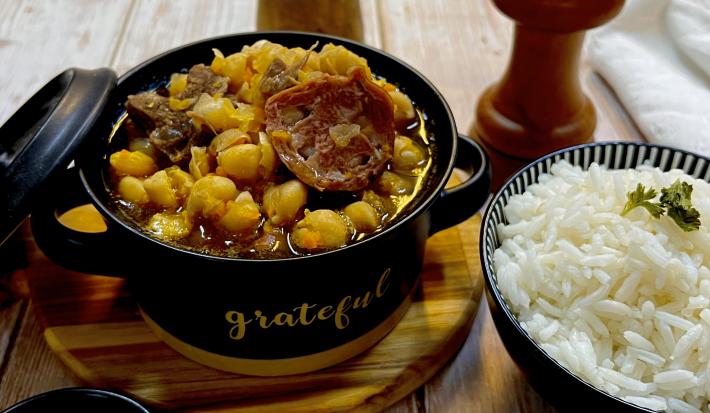 Foto da receita de Grão de Bico com Carnes. Observa-se uma mini panela com grão de bic e linguiça com carne e, ao lado direito, uma mini panela com arroz.