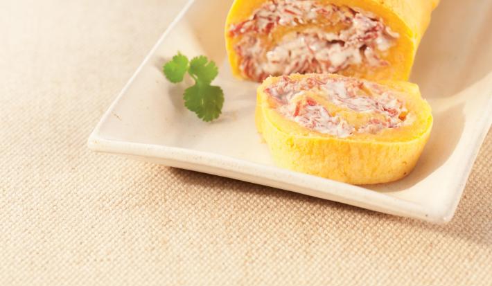 Fotografia em tons de amarelo em uma bancada de madeira com um recipiente retangular e o rocambole de mandioca com carne-seca cortado ao meio.